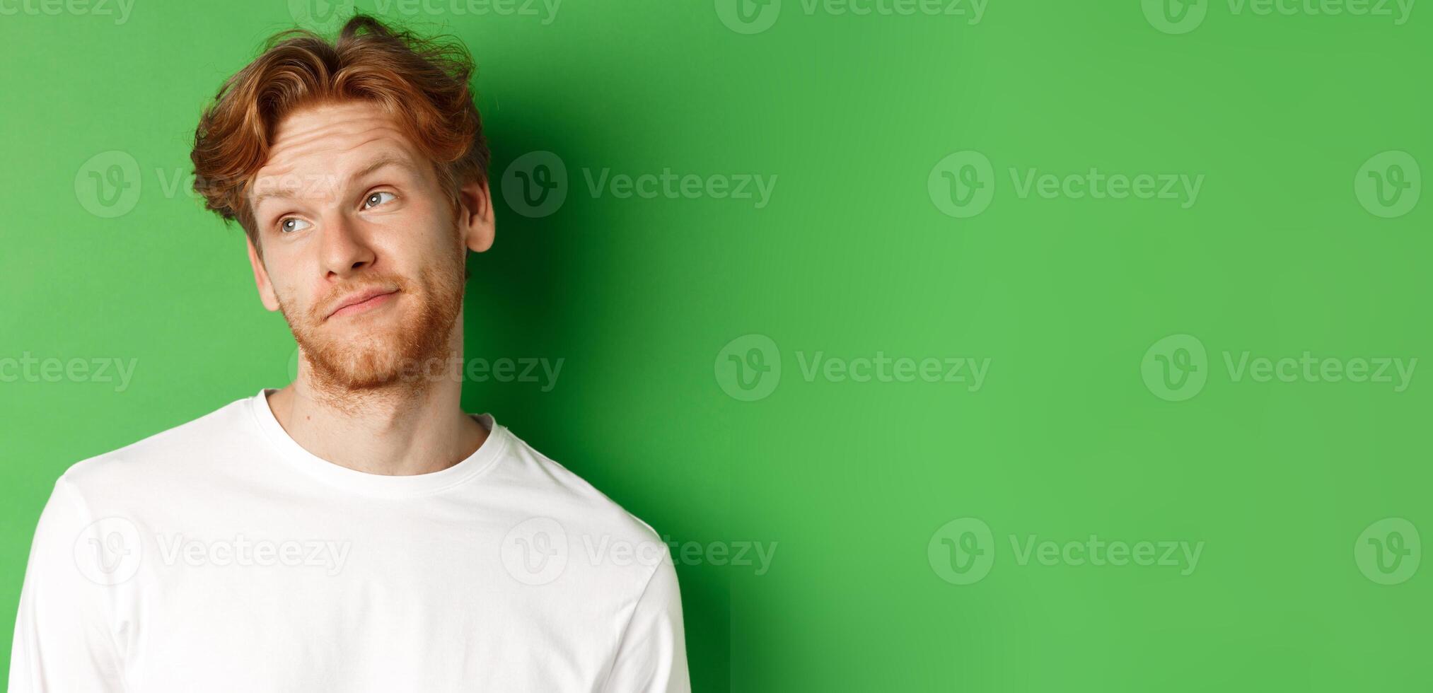 Smiling redhead man with messy haircut and beard tilt head, looking left with pleased face, standing over green background photo