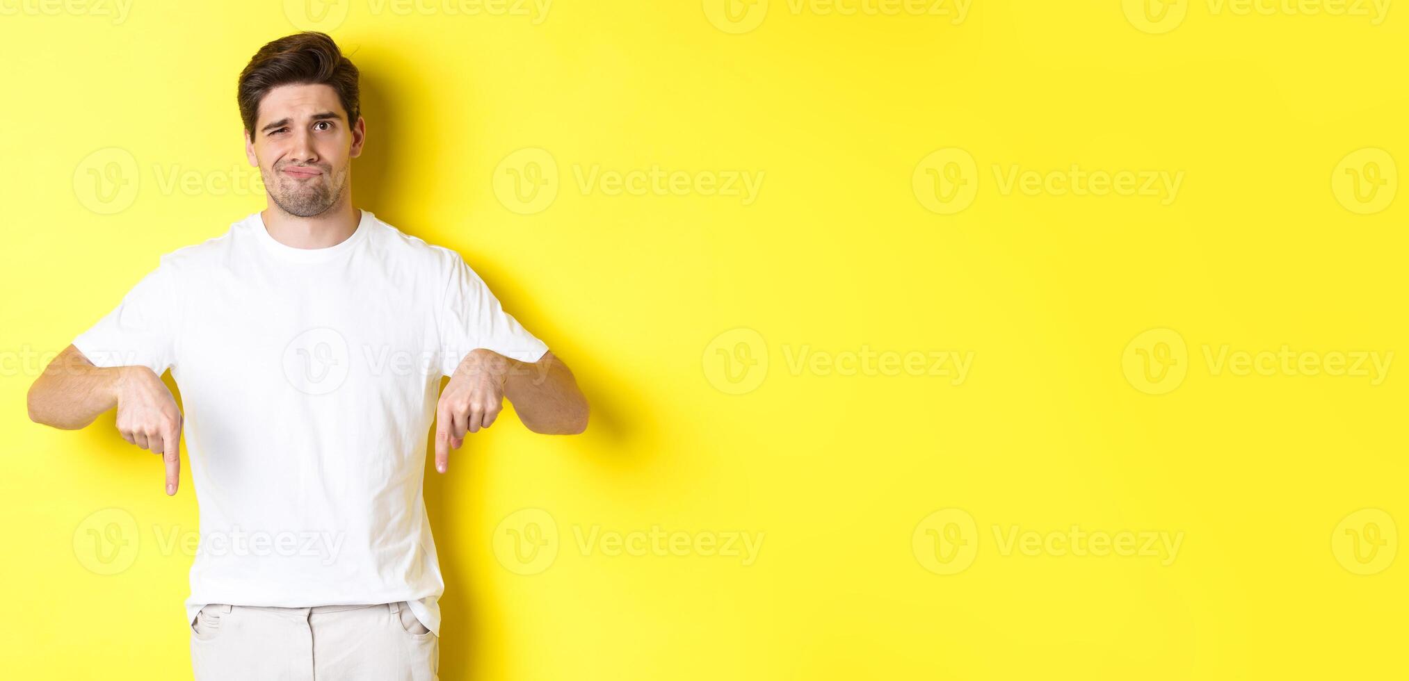 Young unhappy guy grimacing, pointing fingers down at advertisement, disappointed in product, standing over yellow background photo