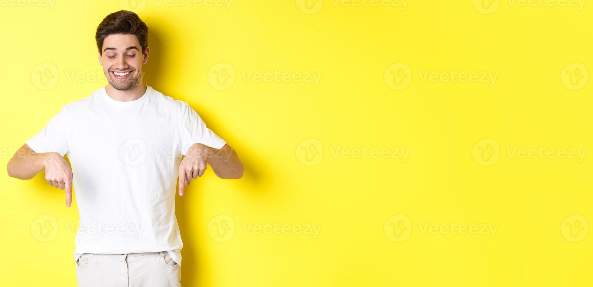 Young handsome man pointing and looking down, checking out promo offer, standing over yellow background photo