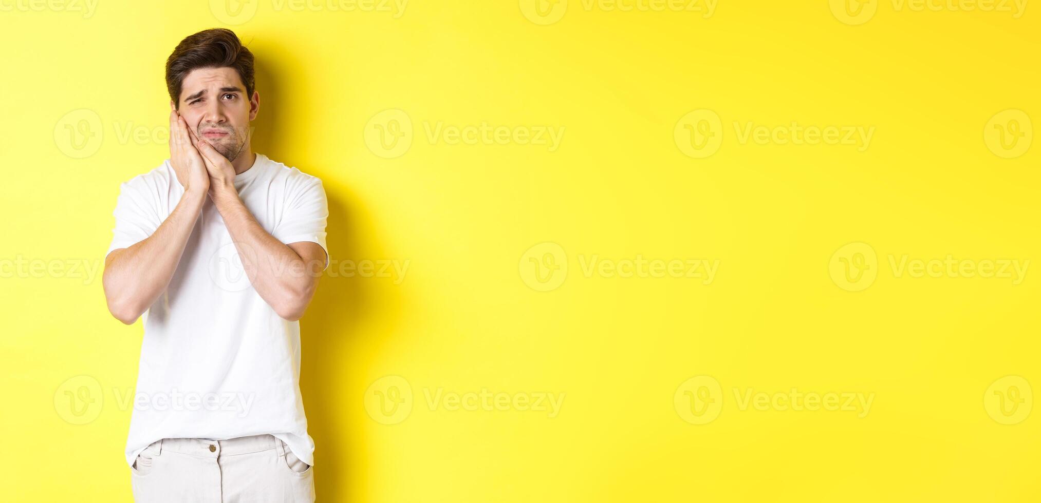 Sad guy complaining on toothache, touching swallen cheek and looking upset, standing over yellow background photo