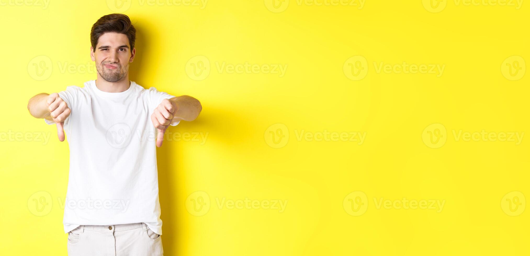 Skeptical guy showing thumbs down and smirking, dislike and disagree, standing over yellow background photo