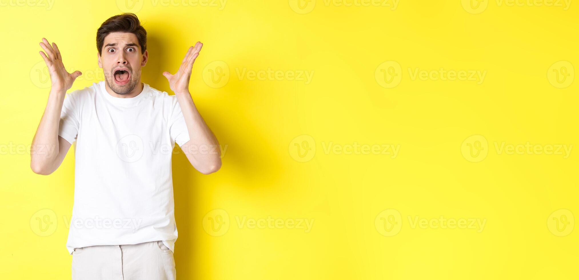 Frustrated and shocked guy panicking, screaming and looking scared, standing in white t-shirt over yellow background photo