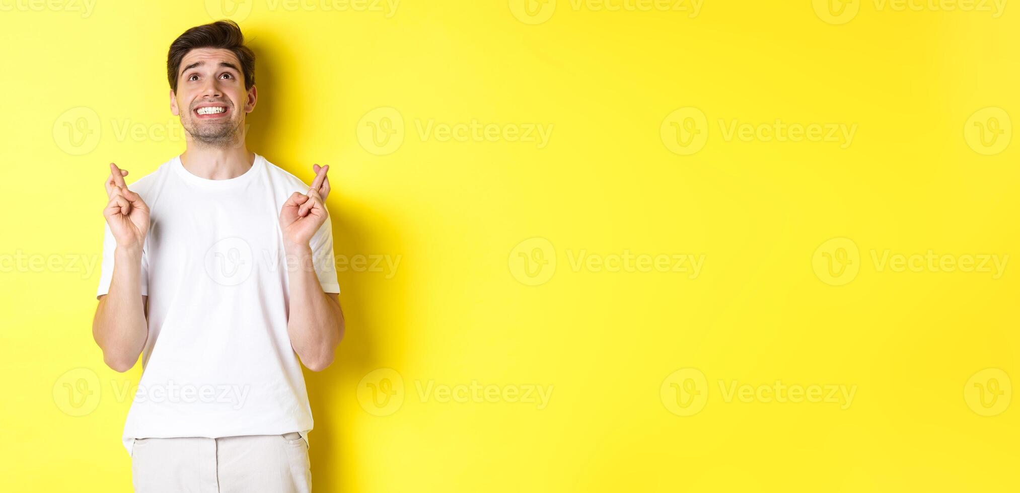 Nervous and hopeful man praying to god, making wish with fingers crossed, panicking and standing over yellow background photo