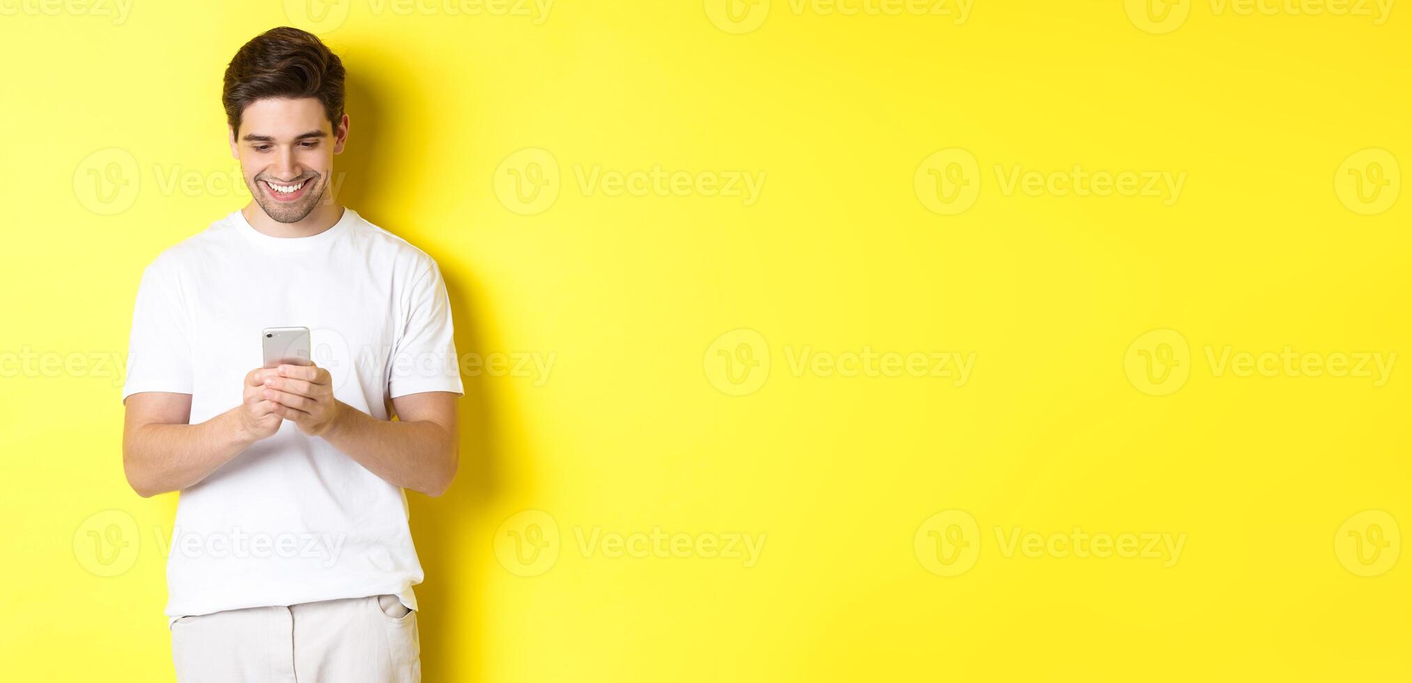 Young man reading text message on smartphone, looking at mobile phone screen and smiling, standing in white t-shirt against yellow background photo