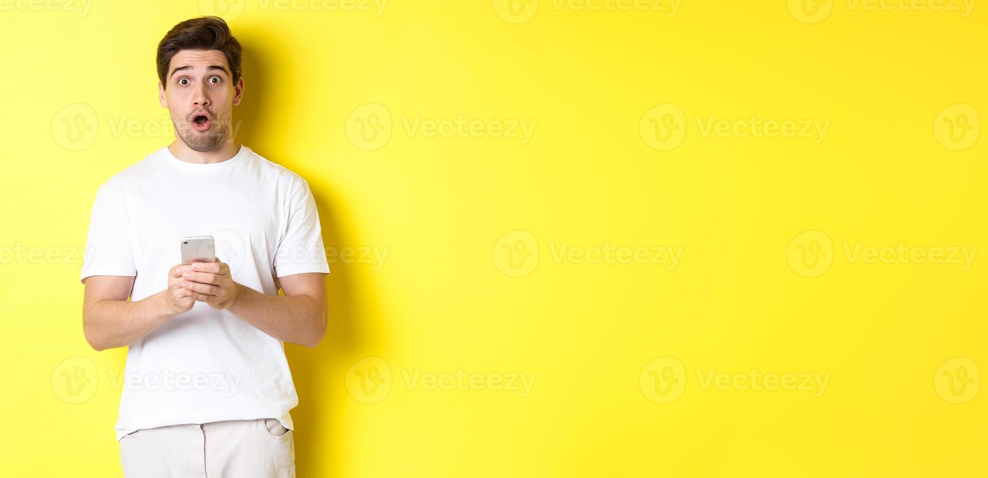 Man looking surprised, using smartphone, open mouth and saying wow, standing against yellow background photo