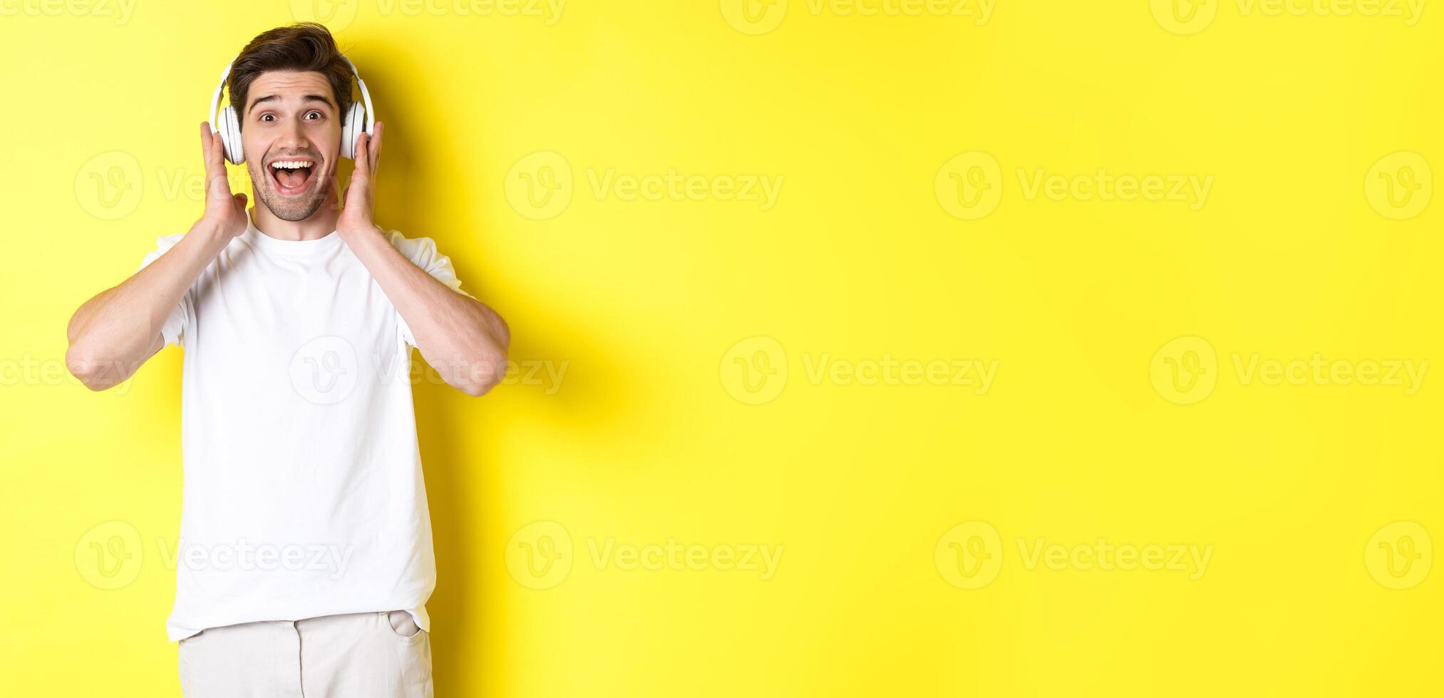 Man in headphones looking surprised and happy, listening awesome song, standing over yellow background photo