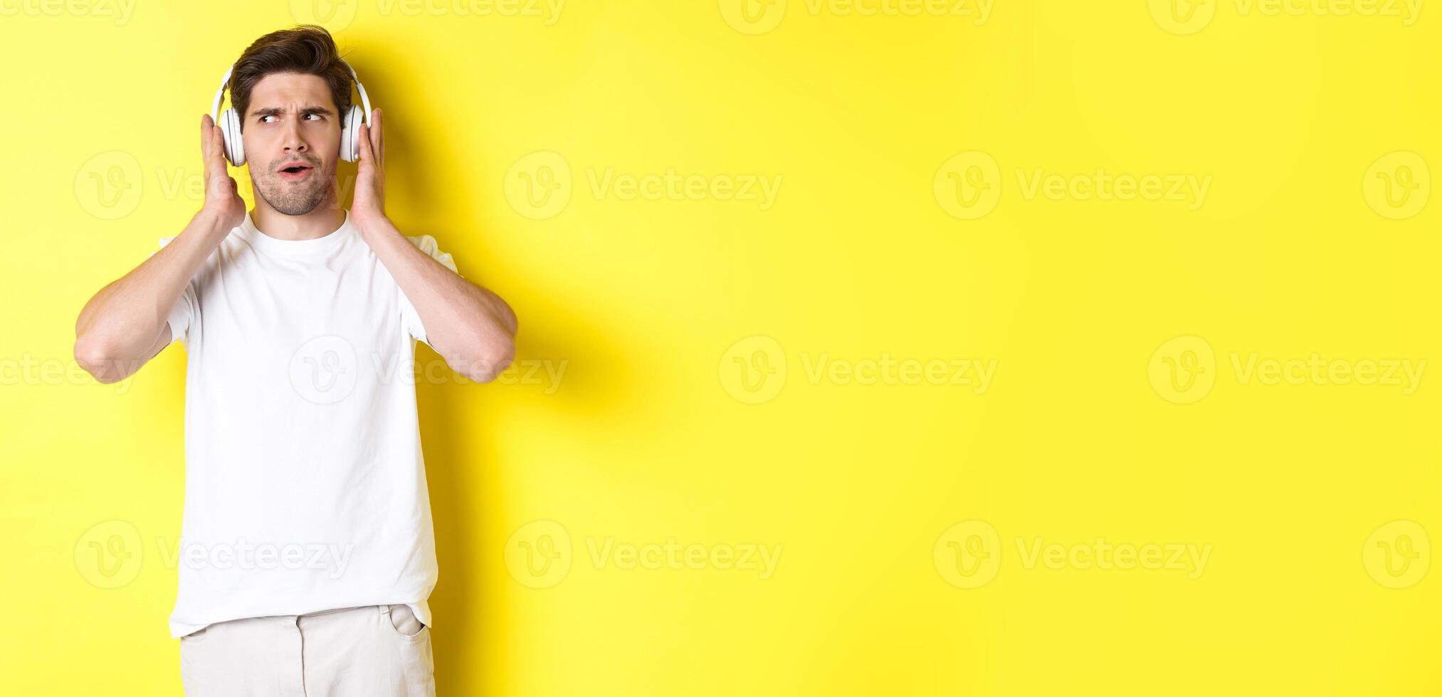 Intrigued guy enjoying tunes in headphones, listening closely to music in earphones, standing over yellow background photo