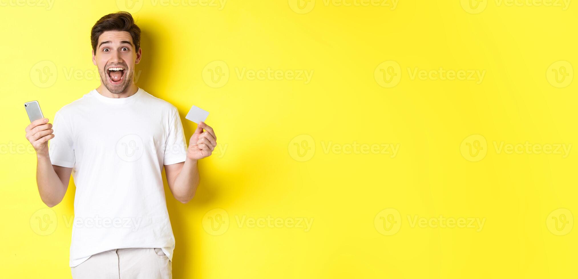 Surprised guy holding smartphone and credit card, online shopping on black friday, standing over yellow background photo