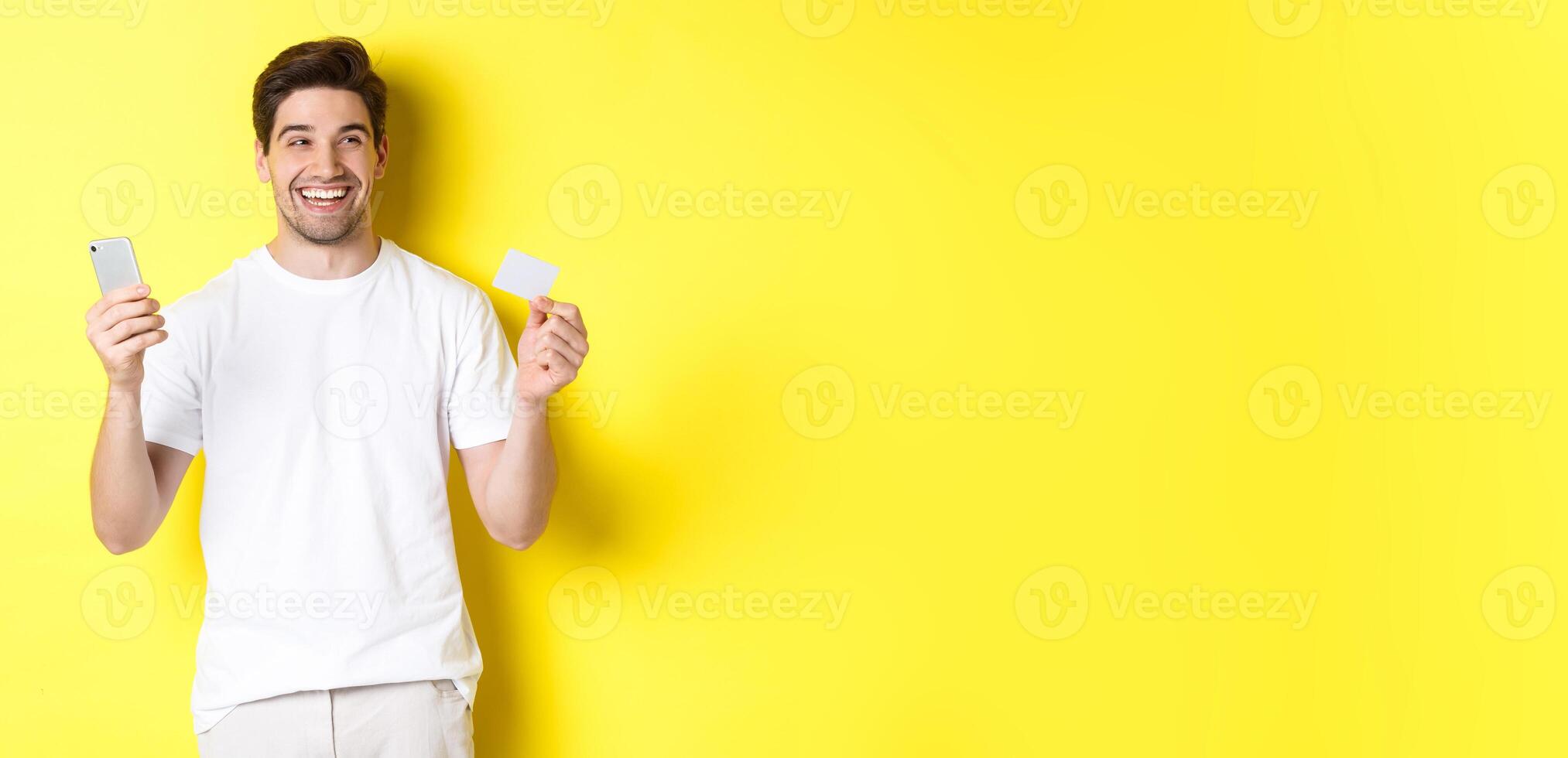 Happy man thinking about shopping, holding credit card and smartphone, smiling pleased, standing over yellow background photo