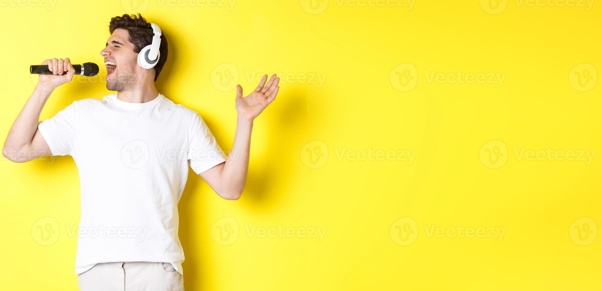 Passionate guy in headphones holding microphone, singing karaoke song, standing over yellow background in white clothes photo