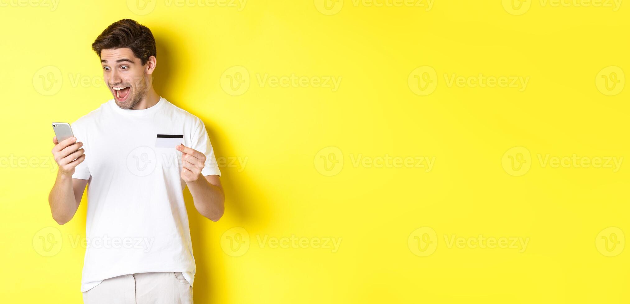 Man looking surprised at smartphone, shopping online, holding credit card, standing over yellow background photo