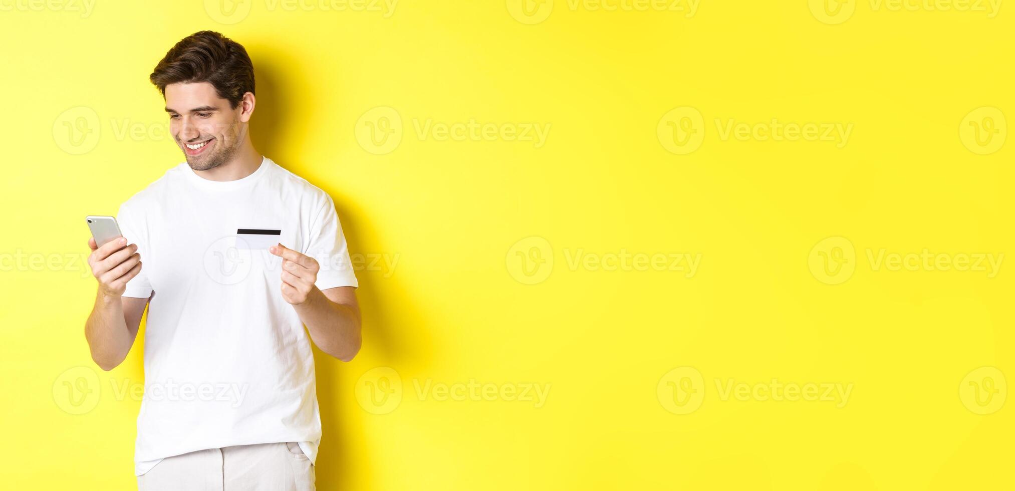 Young man paying online, insert credit card number on mobile phone, shopping in internet, standing over yellow background photo