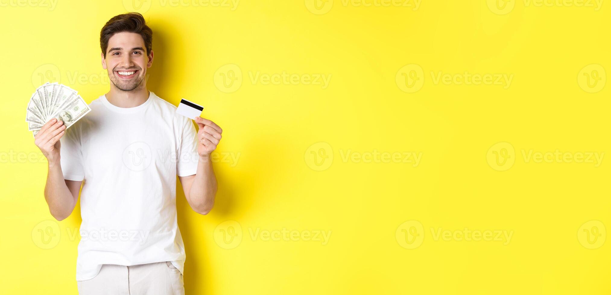 Young man withdraw money from credit card, smiling pleased, standing over yellow background photo