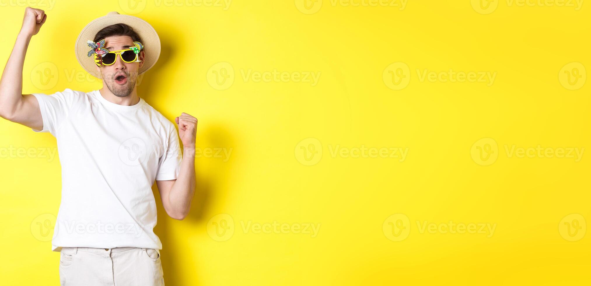 Concept of tourism and lifestyle. Happy guy tourist enjoying trip, rooting for you, fist pump and triumphing, going on journey in summer hat and sunglasses, yellow background photo