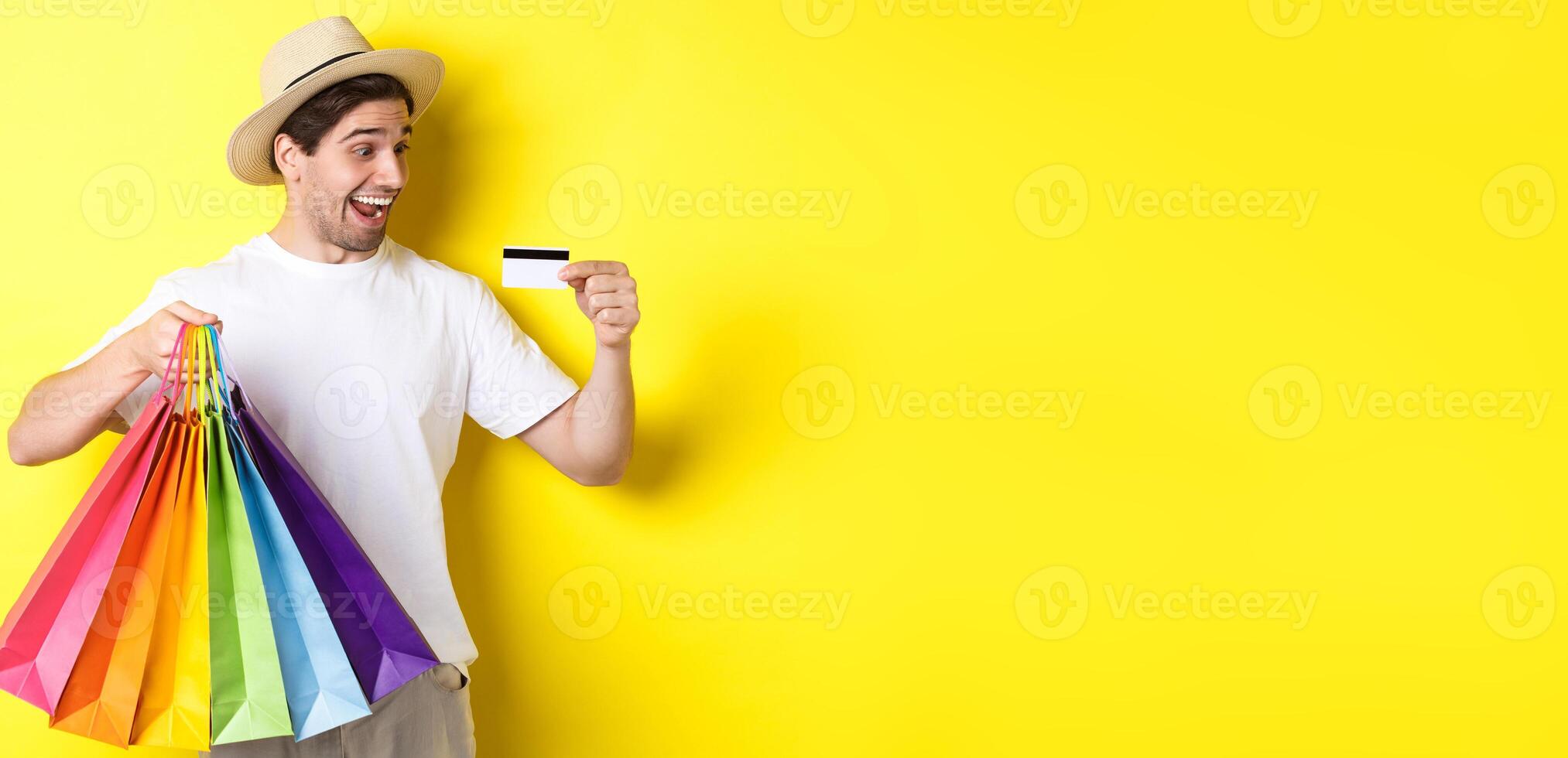 Excited man going shopping on vacation, looking satisfied at credit card, holding paper bags, standing over yellow background photo