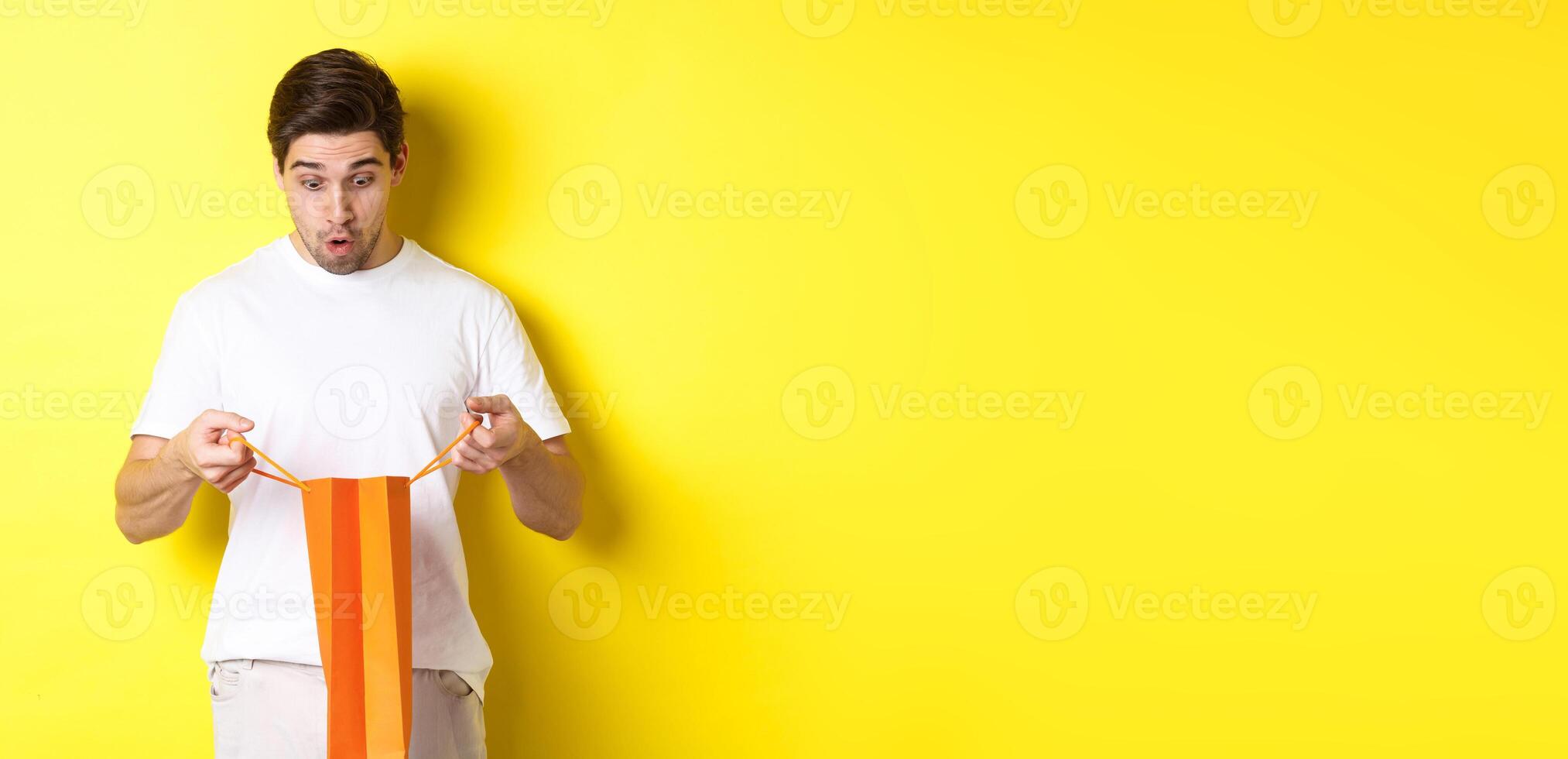 Surprised boyfriend open bag with gift, looking inside amazed, standing over yellow background photo
