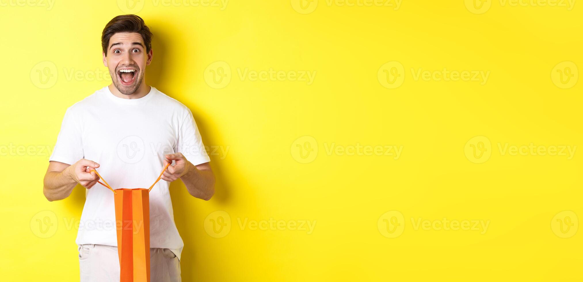 Surprised guy open shopping bag with fist, looking excited and happy at camera, standing against yellow background photo