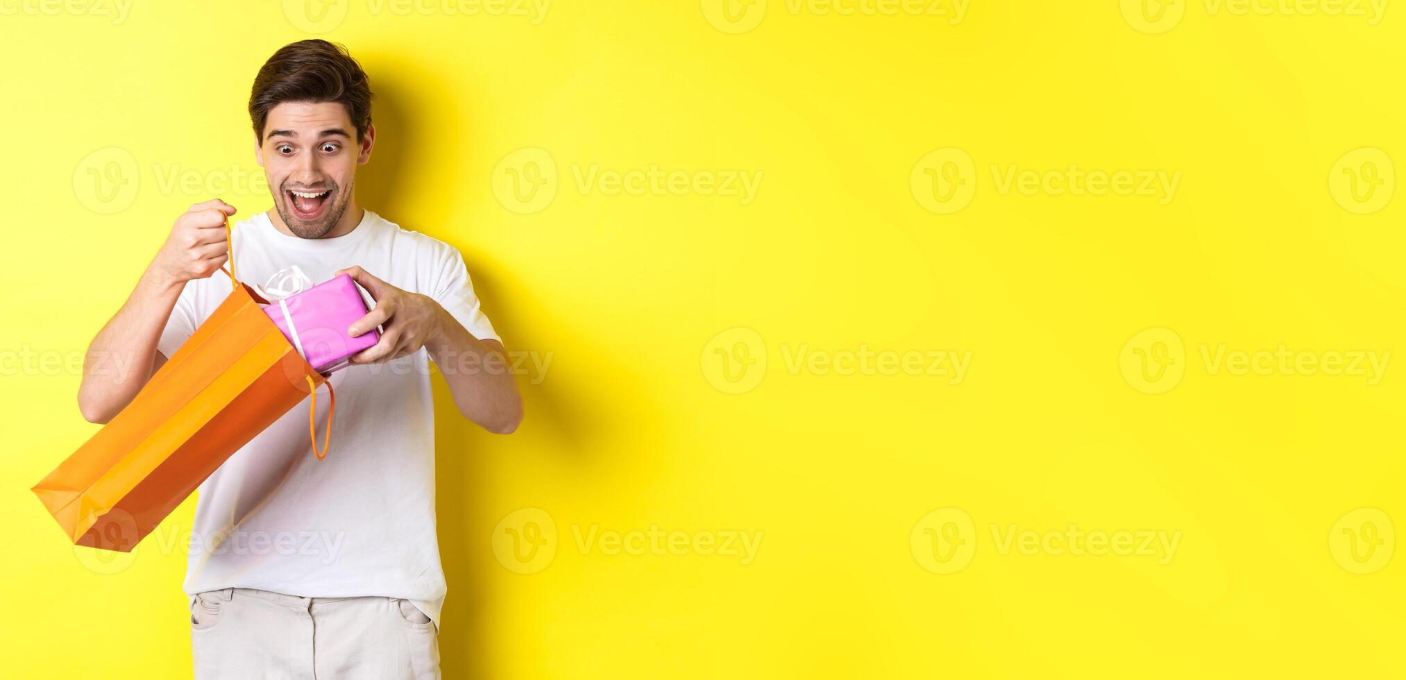 Concept of holidays and celebration. Young man looking surprised as take out gift from shopping bag, standing over yellow background photo