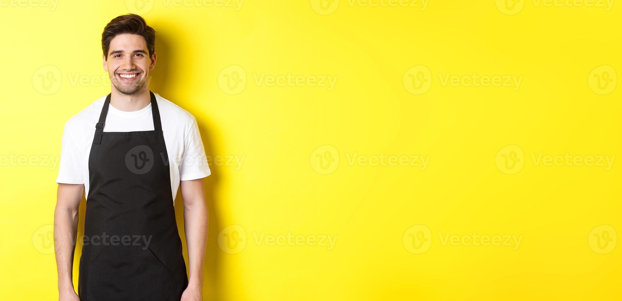 Young man waiter in black apron, smiling, working in store or coffee shop, standing against yellow background photo