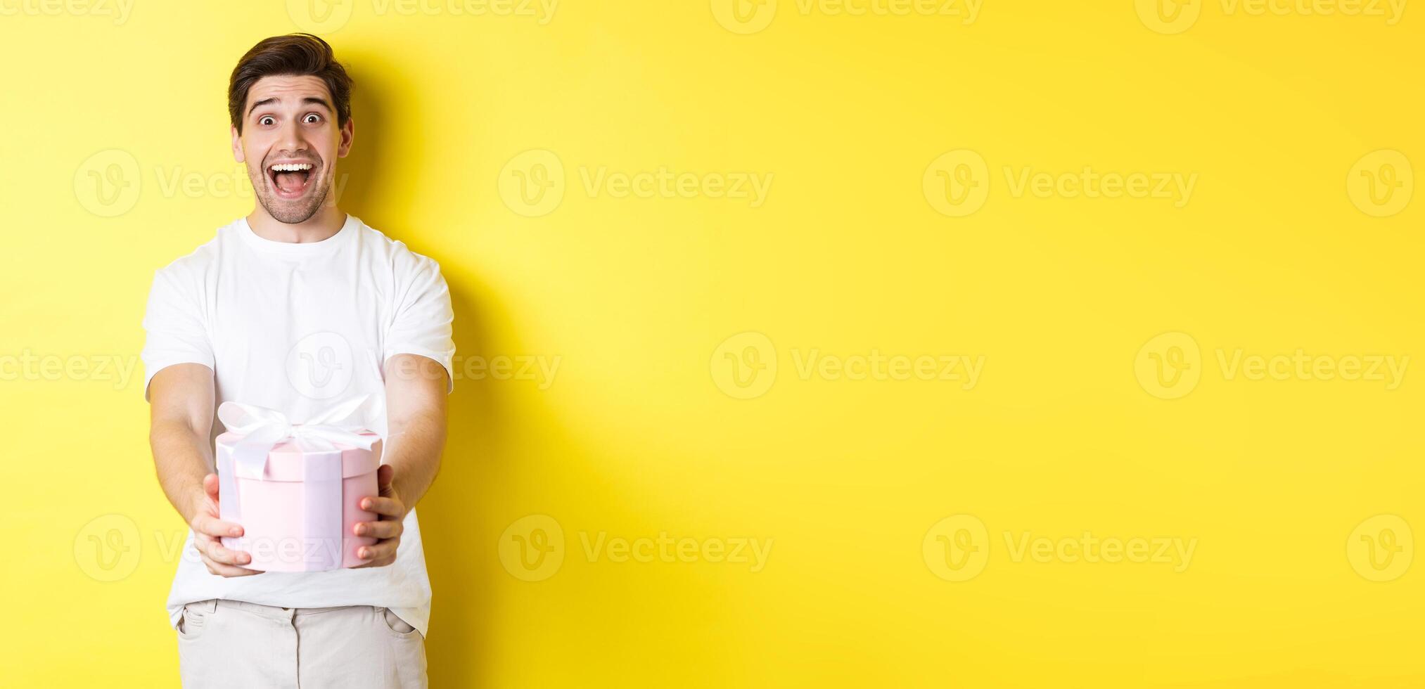 Concept of holidays and celebration. Happy man giving a gift and looking excited, standing against yellow background photo