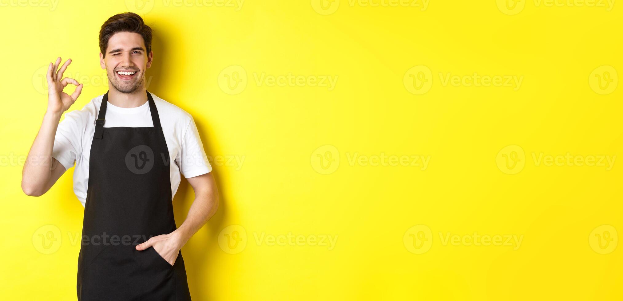 Confident and handsome waiter showing ok sign, wearing black apron and standing against yellow background photo