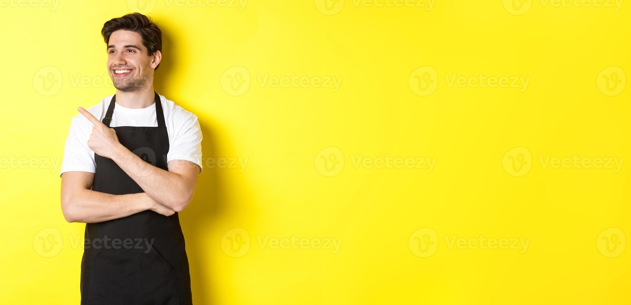 Handsome barista pointing and looking left at promo, wearing black apron, standing against yellow background photo