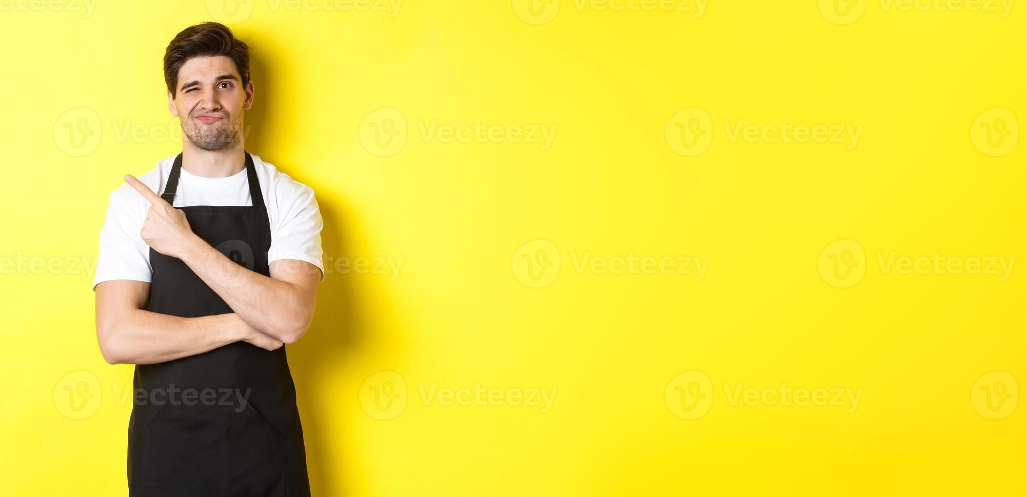 Displeased barista in black apron pointing finger at upper left corner, grimacing skeptical, dislike something, standing against yellow background photo