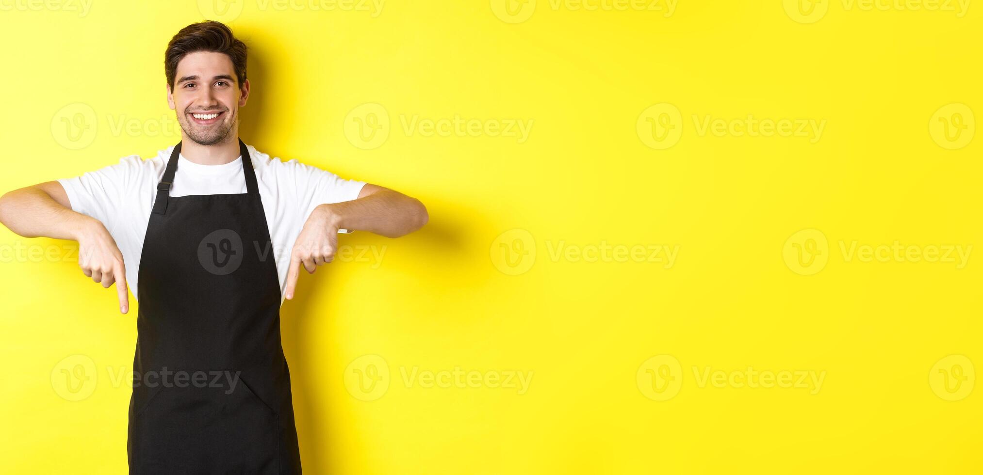 Friendly barista in black apron pointing fingers down, showing your logo banner, standing over yellow background photo