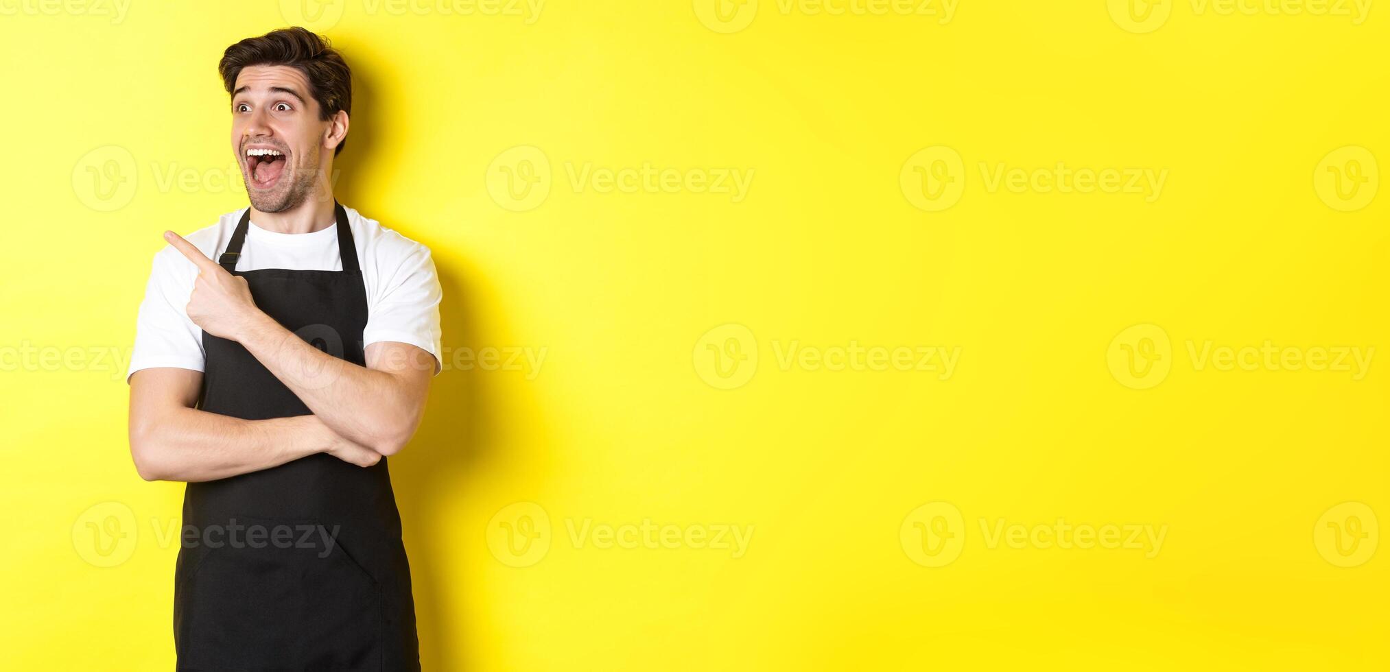 Waiter in black apron checking out promo offer, pointing finger and looking left at your logo, standing over yellow background photo