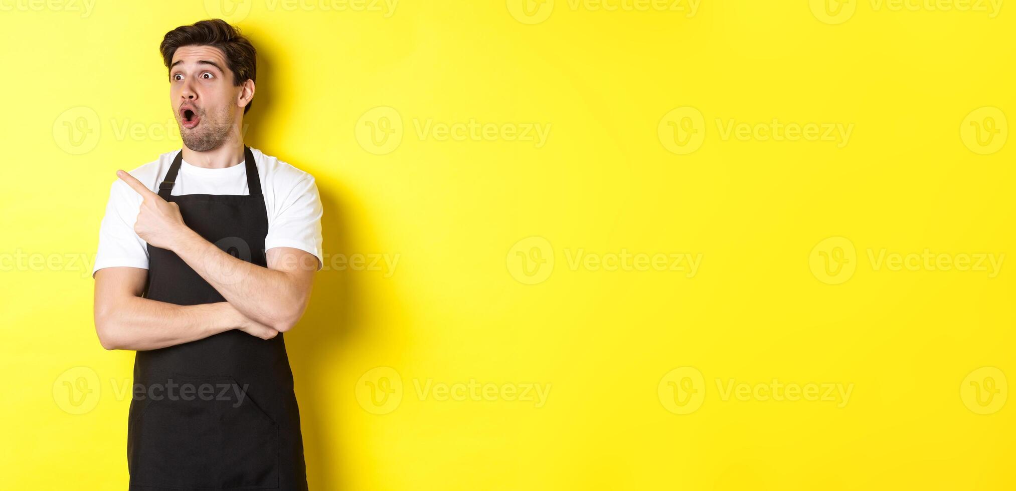 Waiter in black apron checking out promo offer, pointing finger and looking left at your logo, standing over yellow background photo