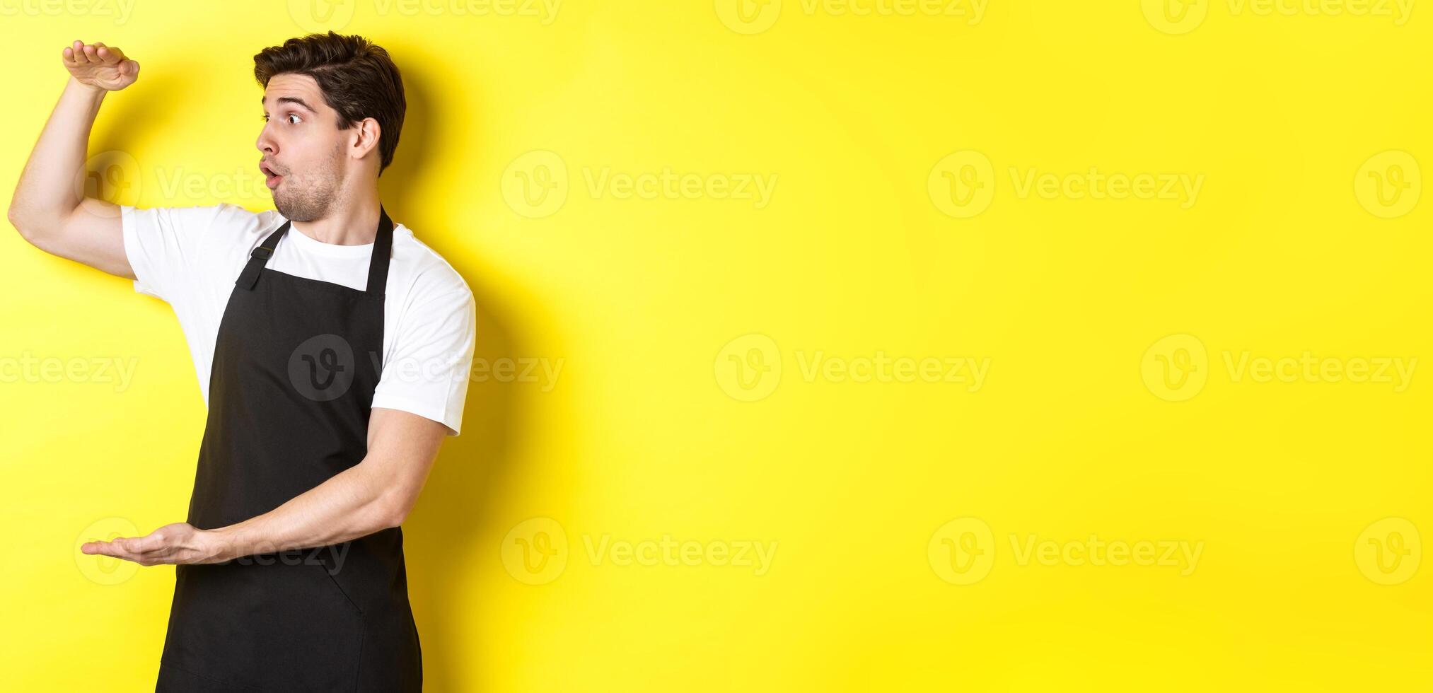 Waiter in black apron looking amazed at something large, holding big object, standing over yellow background photo
