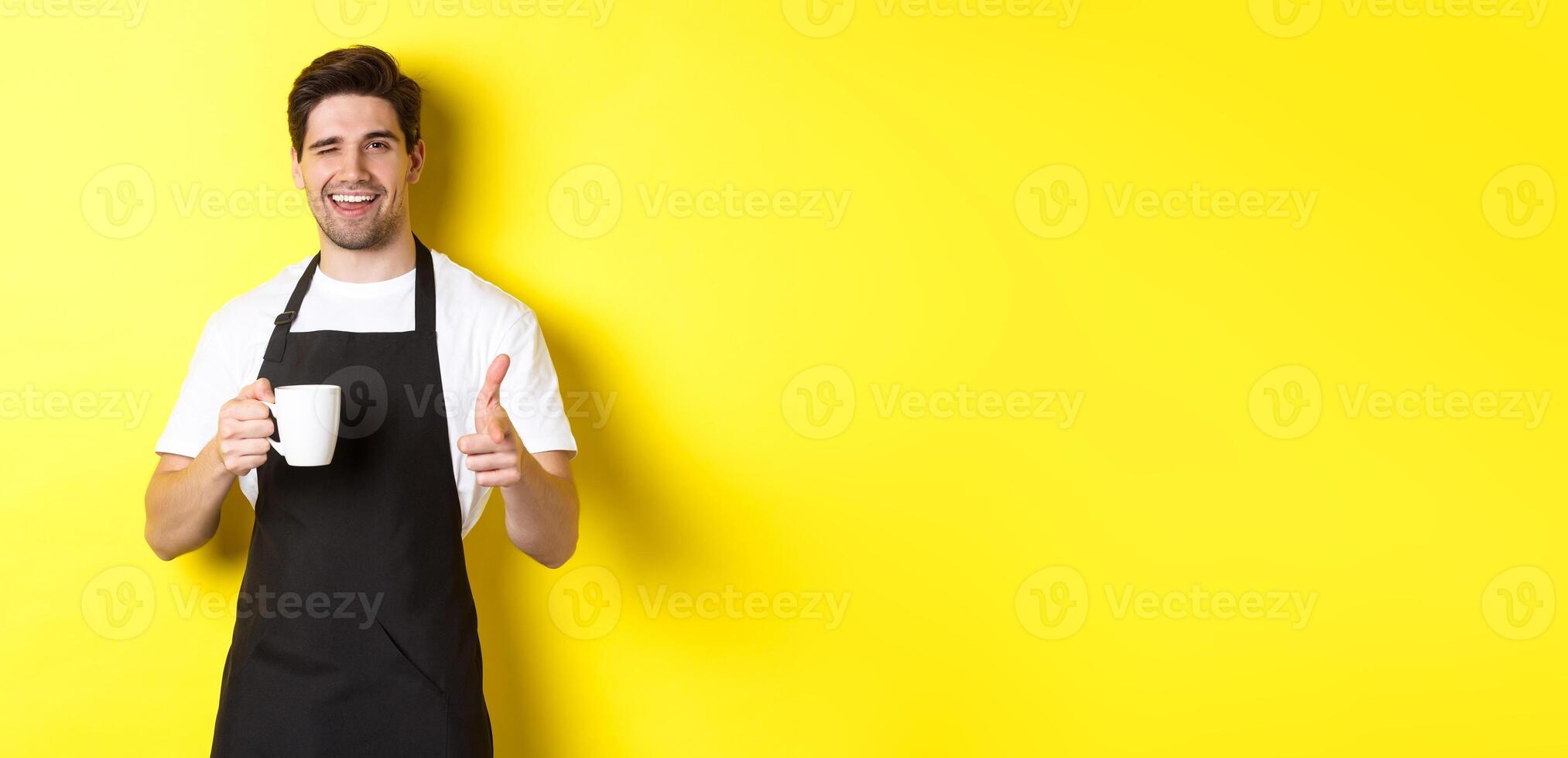 Handsome barista in black apron holding coffee cup, pointing finger at you, inviting visit cafe, standing over yellow background photo