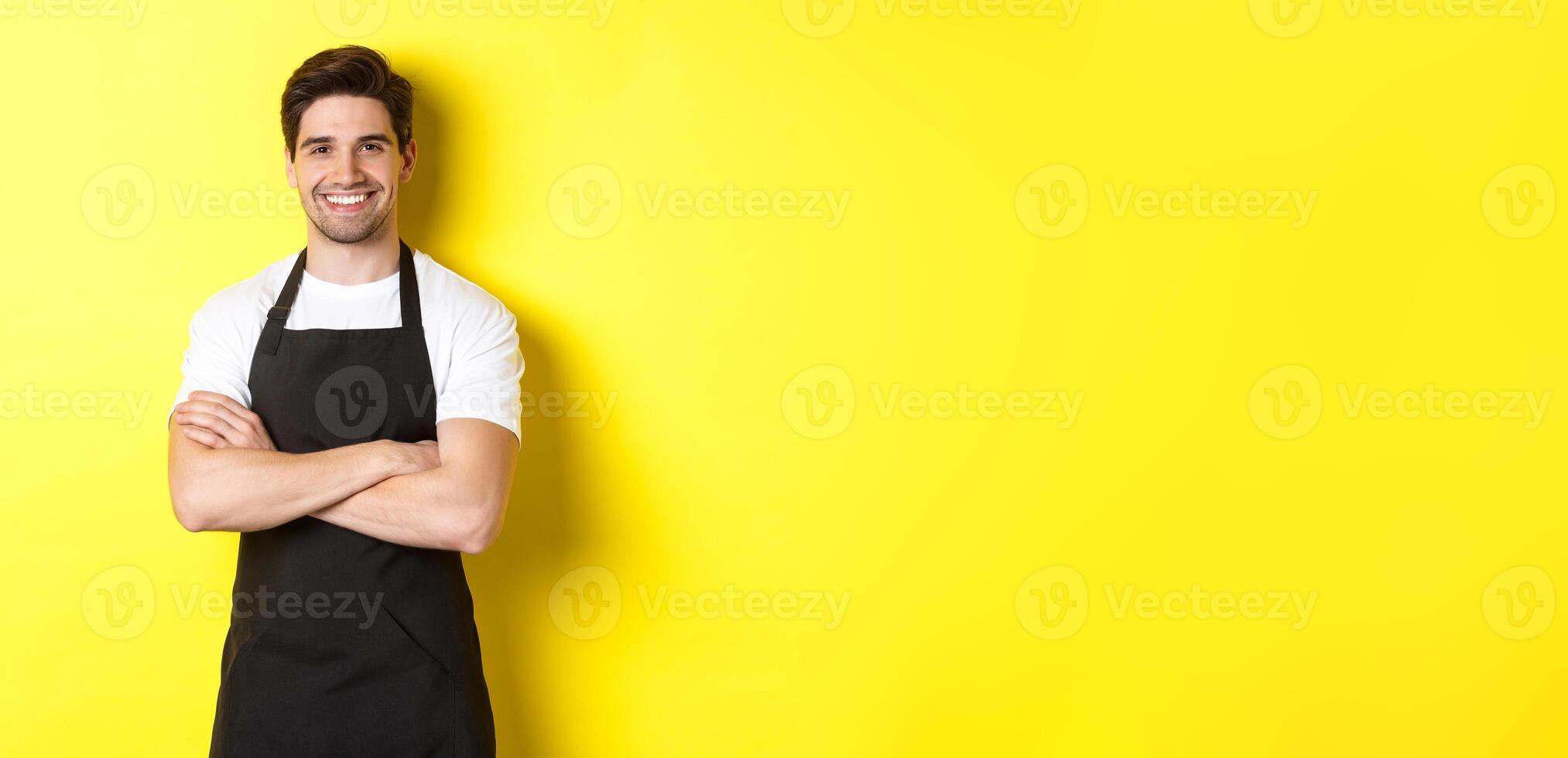 Smiling male waiter in black apron standing confident, cross arms on chest against yellow background photo