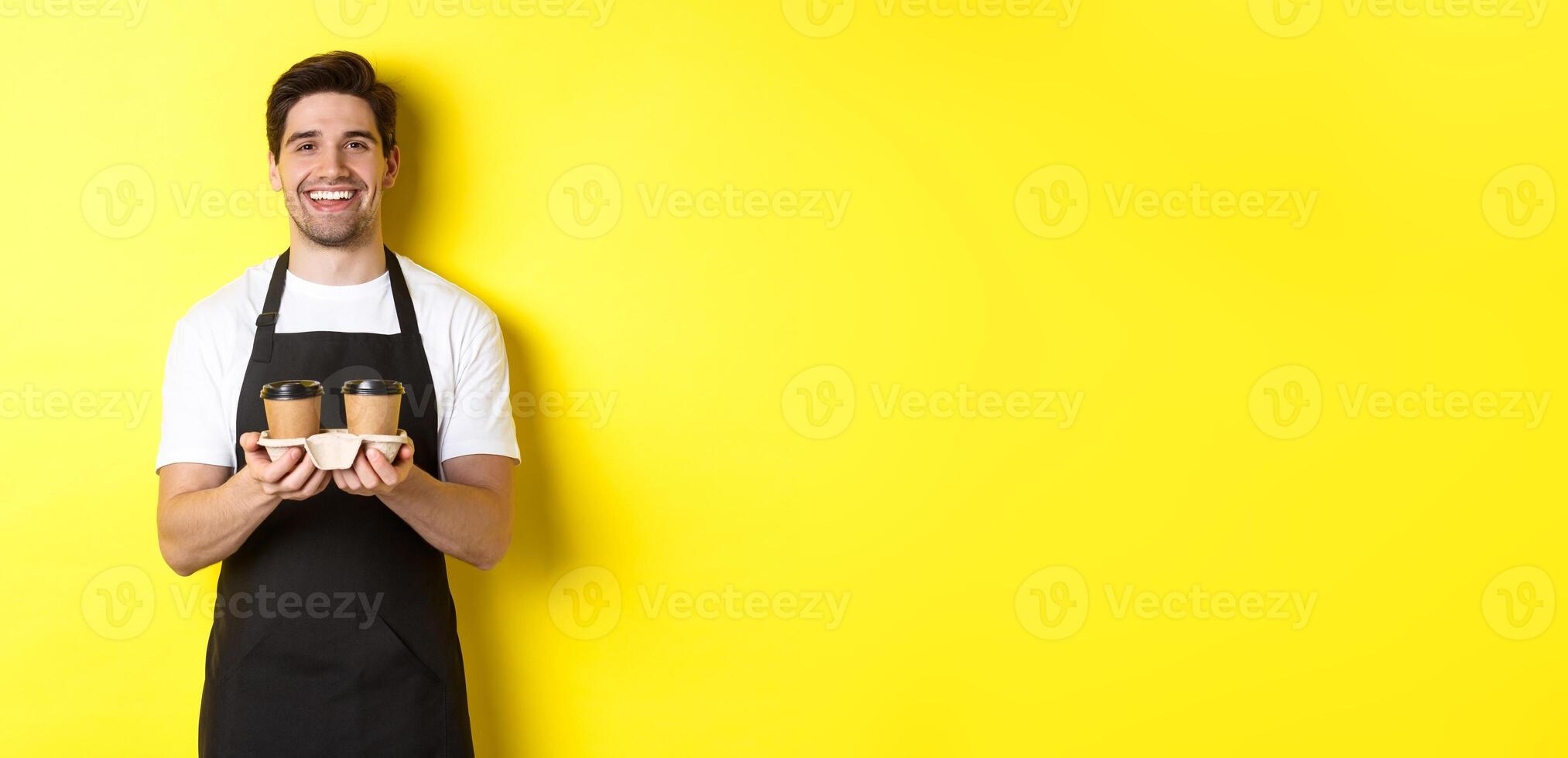 Friendly barista in black apron giving takeaway order, holding two cups of coffee and smiling, standing over yellow background photo