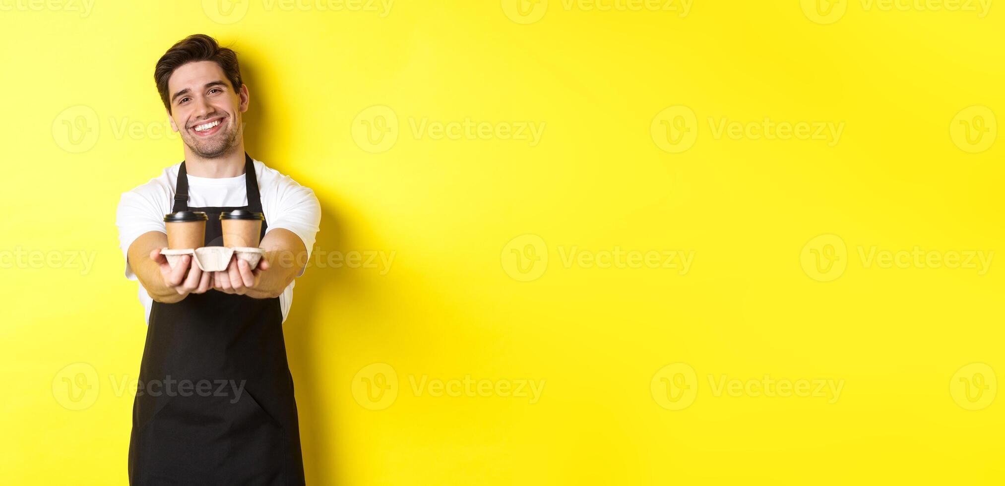 Friendly barista in black apron giving takeaway order, holding two cups of coffee and smiling, standing over yellow background photo