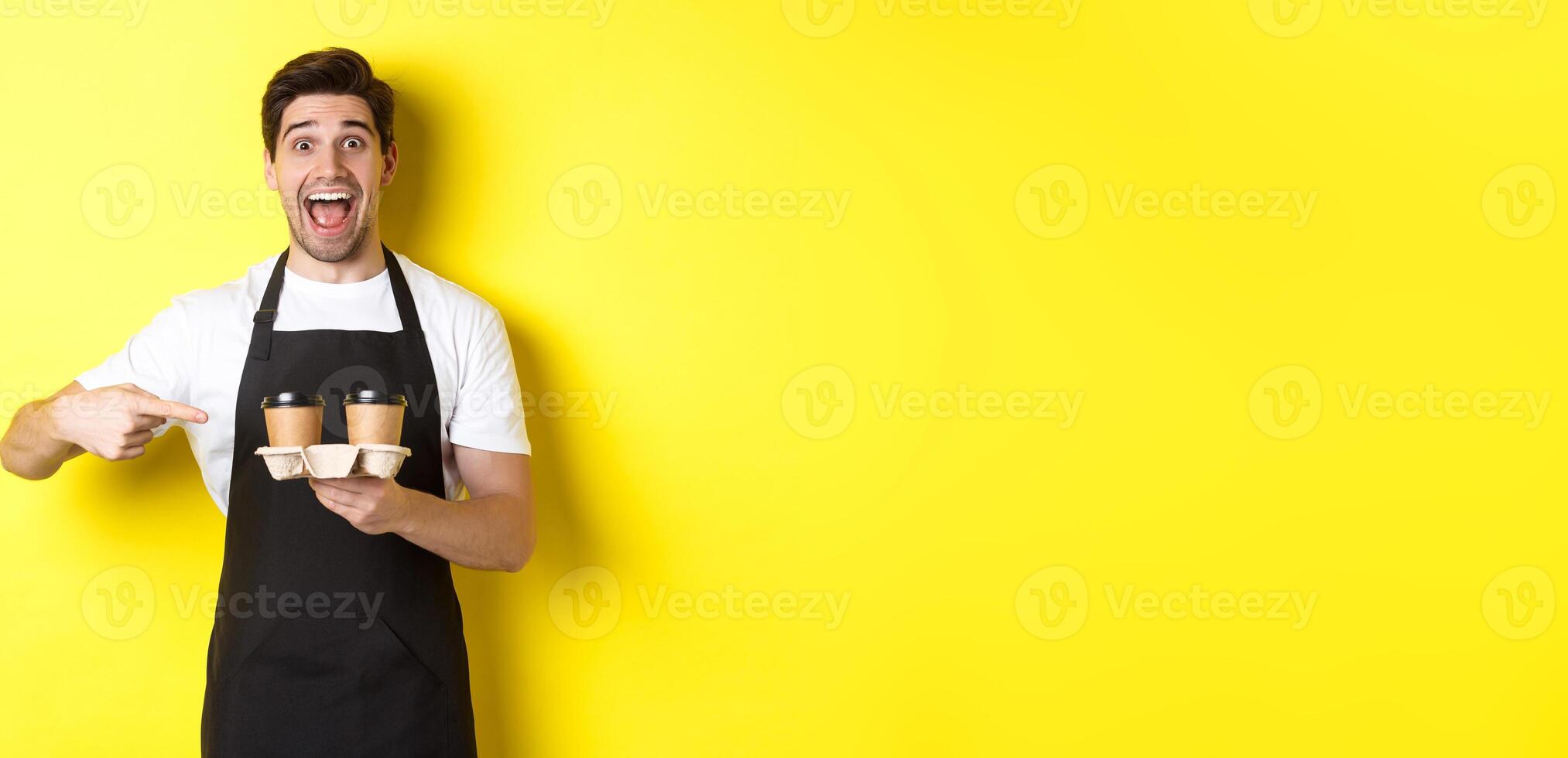 Excited barista in black apron pointing fingers at takeaway coffee cups, standing against yellow background happy photo