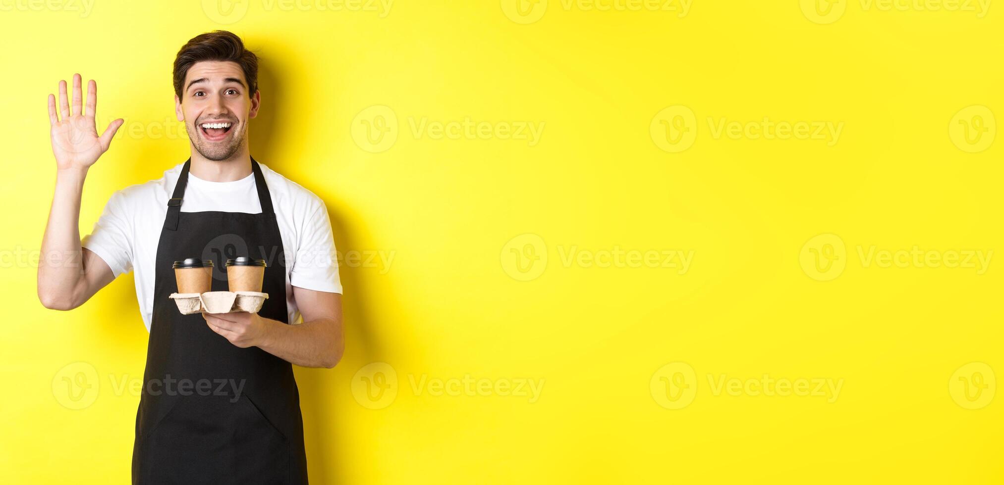 Friendly waiter in cafe waving hand at customer, holding takeaway coffee oder, standing against yellow background in black apron photo