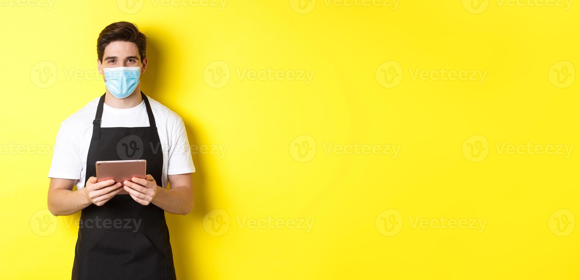 Concept of covid-19, small business and pandemic. Waiter in black apron and medical mask taking order, holding digital tablet, standing over yellow background photo