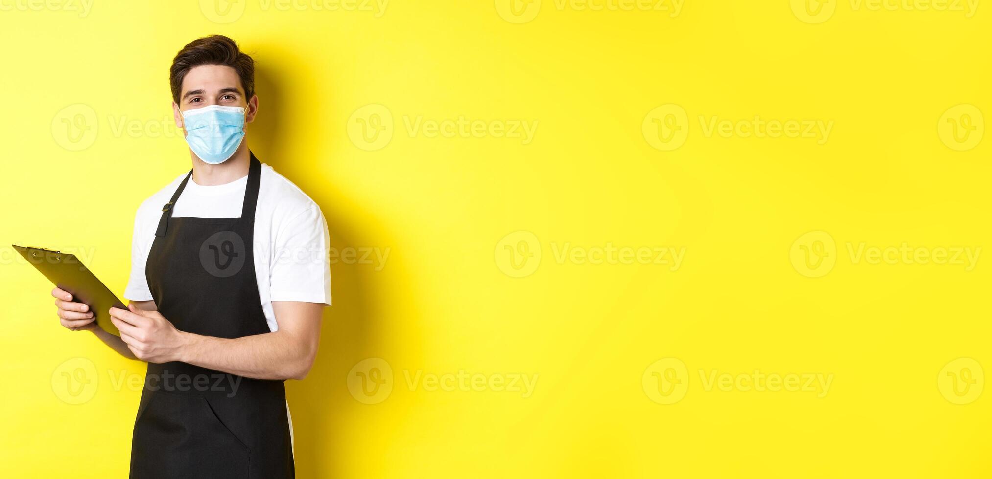Concept of covid-19, small business and quarantine. Young male seller in medical mask and black apron taking orders, holding clipboard, standing over yellow background photo