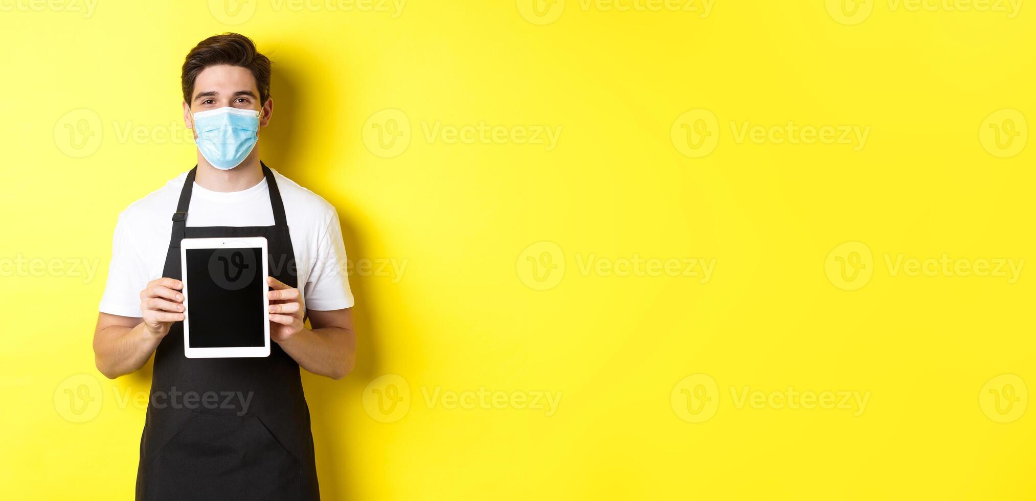 Concept of covid-19, small business and pandemic. Smiling barista in medical mask showing tablet screen, standing over yellow background photo