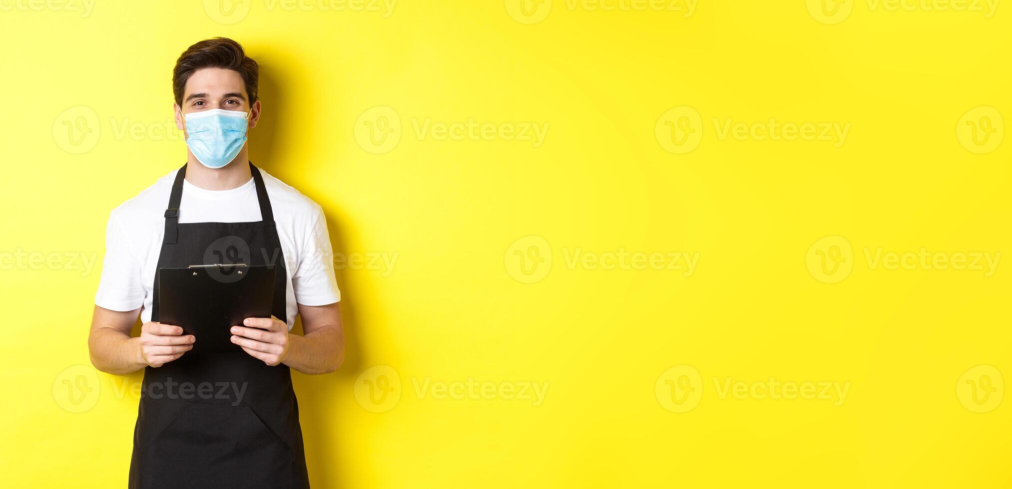 Concept of covid-19, small business and quarantine. Salesman in black apron and medical mask holding clipboard, working in shop, standing over yellow background photo