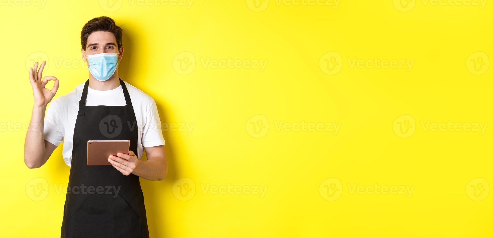 Concept of covid-19, small business and pandemic. Salesman in medical mask and black apron showing ok sign, taking orders with digital tablet, yellow background photo