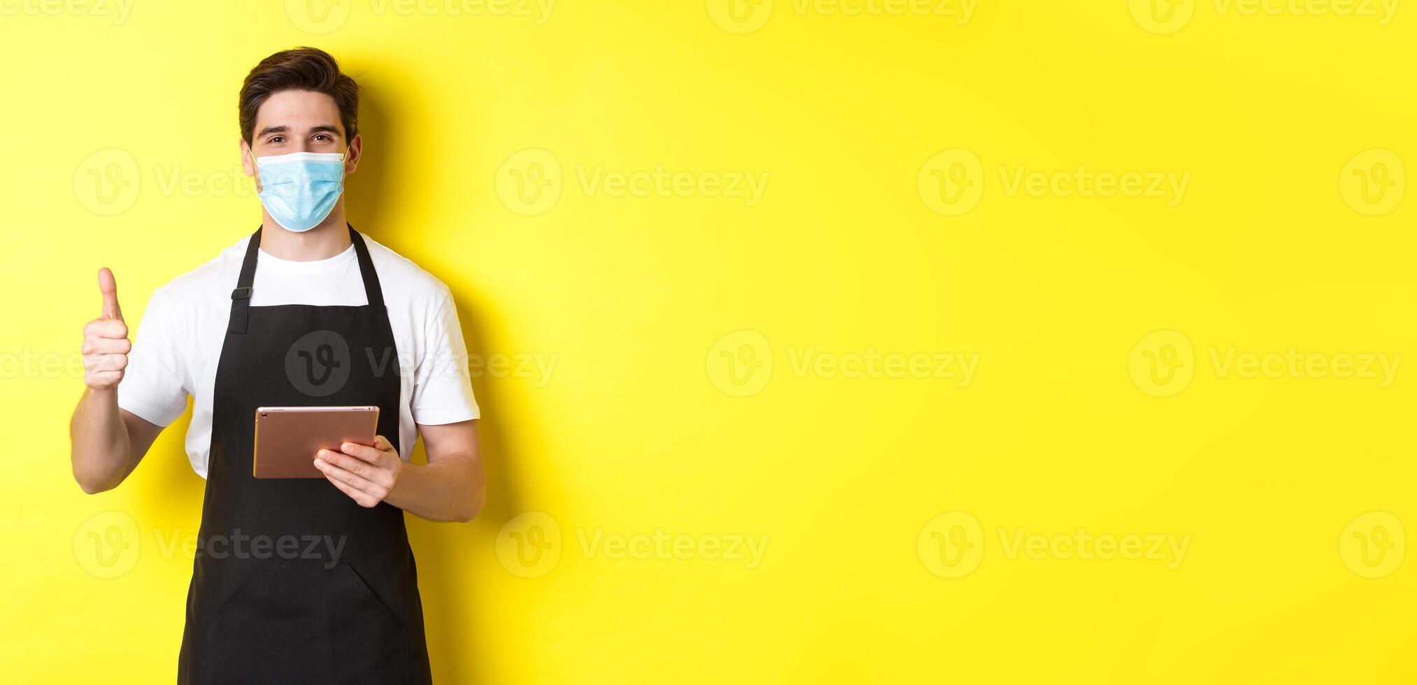 Concept of covid-19, small business and pandemic. Friendly waiter in medical mask and black apron showing thumb up, taking orders with digital tablet, yellow background photo