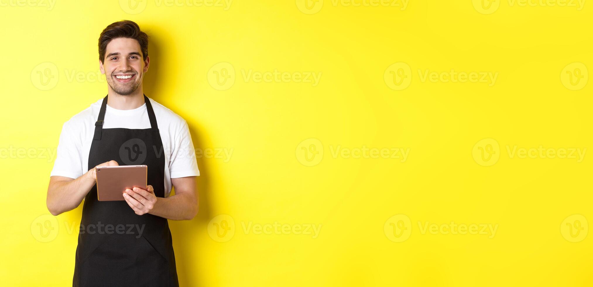 Handsome waiter taking orders, holding digital tablet and smiling, wearing black apron uniform, standing over yellow background photo