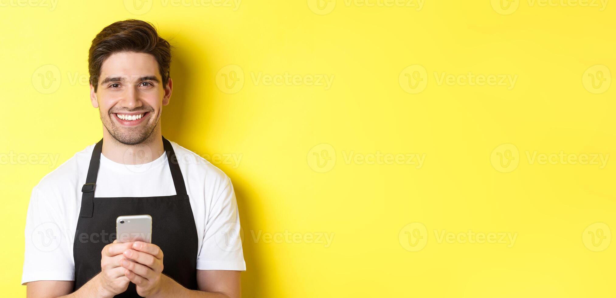 Close-up of handsome barista sending message on mobile phone, smiling happy, standing over yellow background photo