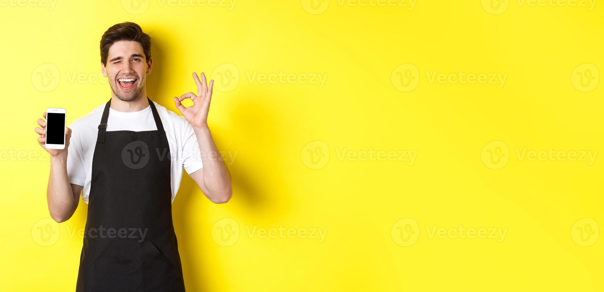 Handsome coffee shop worker showing ok sign and smartphone screen, recommending application, standing over yellow background photo