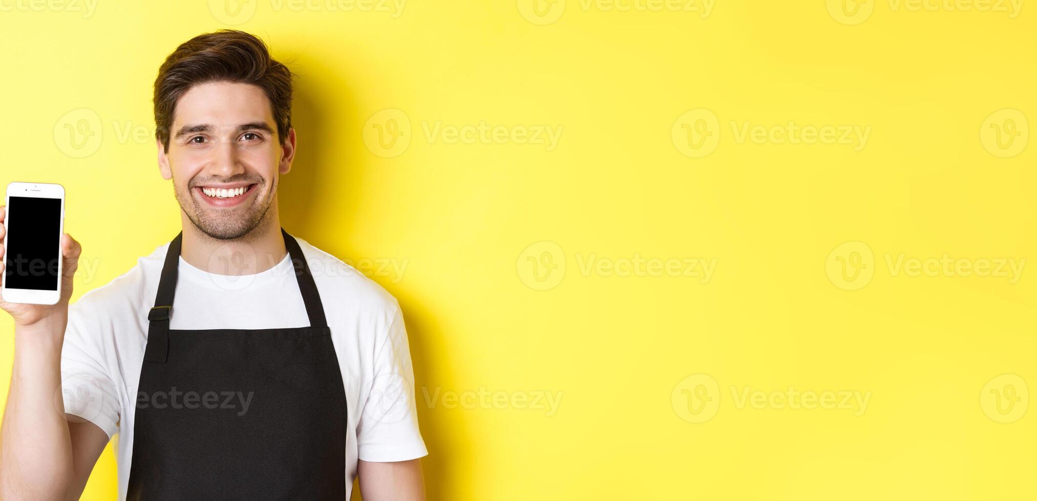 Close-up of handsome waiter in black apron showing smartphone screen, recommending app, standing over yellow background photo