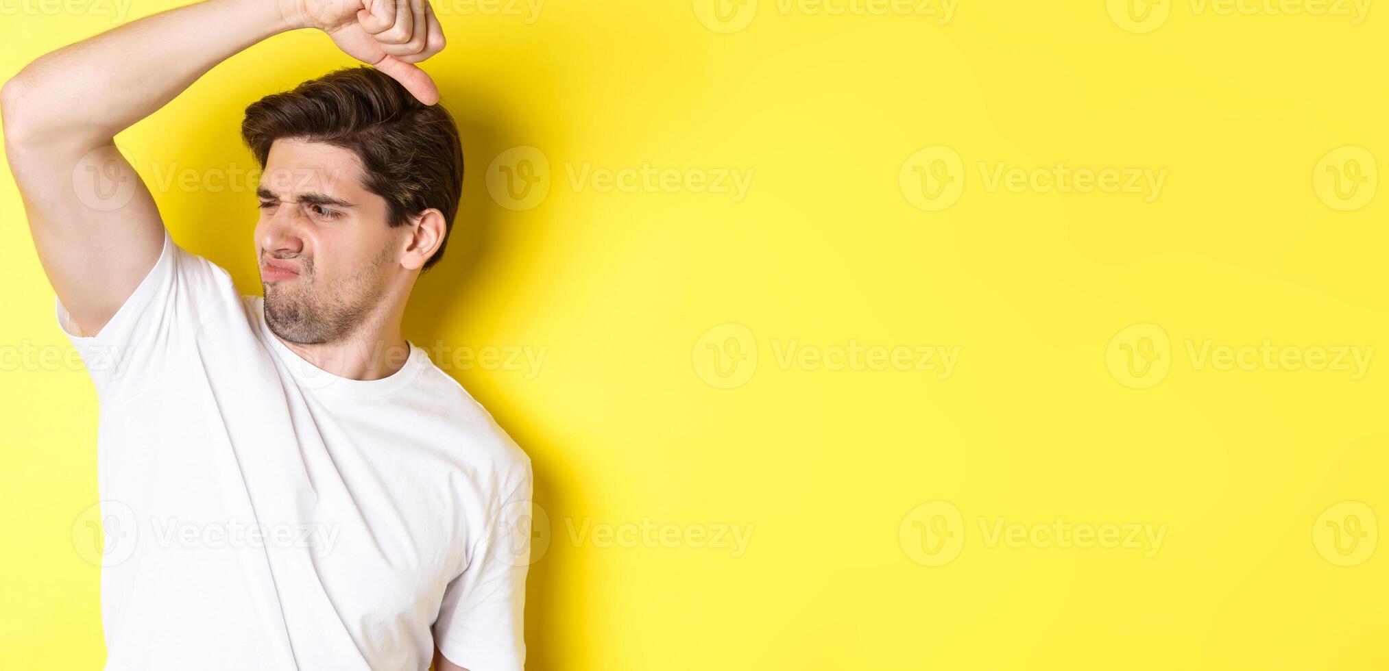 Man in sweat smelling his armpit, standing in white t-shirt and grimacing from stinky clothes photo