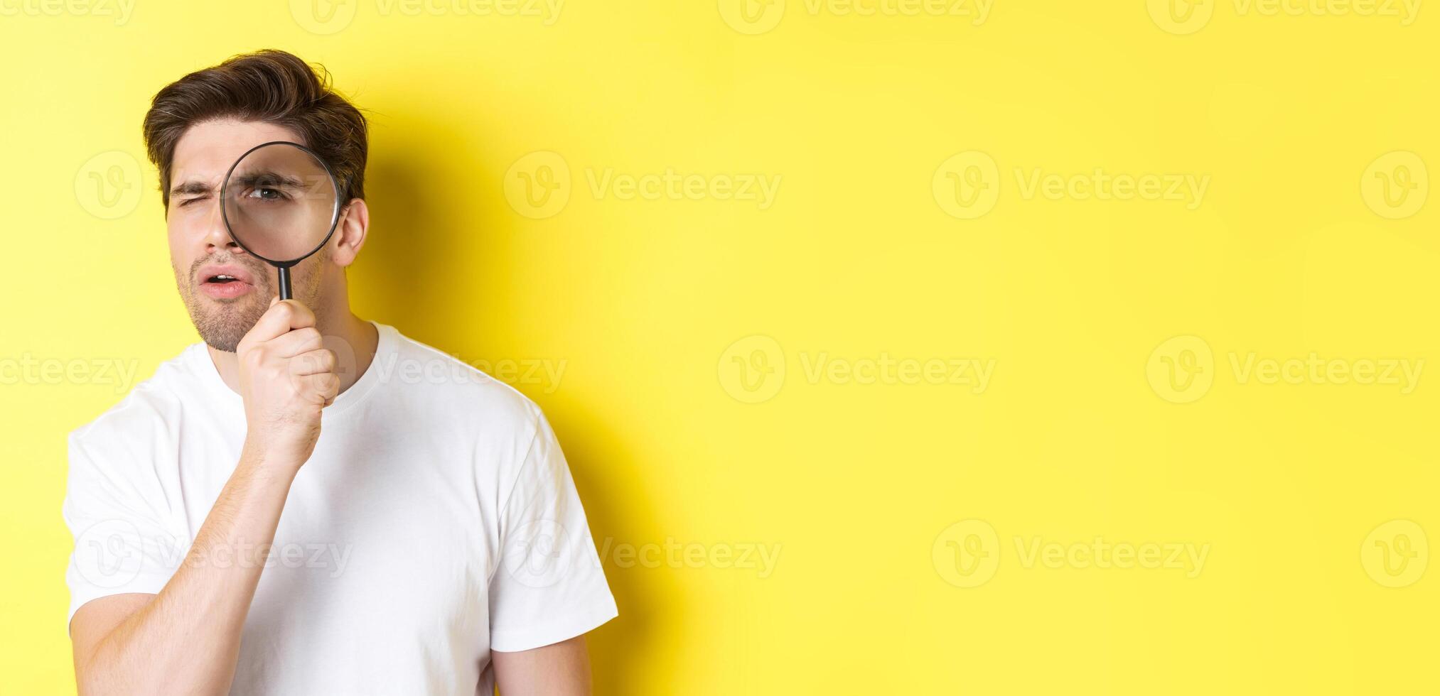 Man looking through magnifying glass with serious and thoughtful look, searching or investigating, standing over yellow background photo
