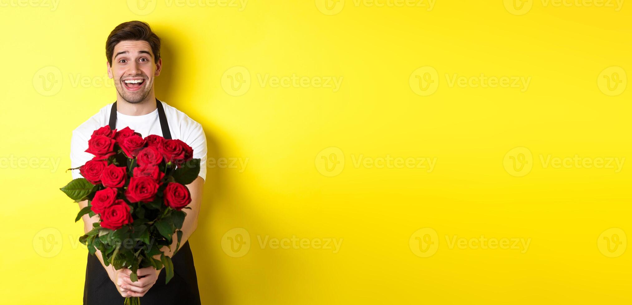 Seller in flower shop wearing black apron, giving bouquet of roses and smiling, standing over yellow background photo